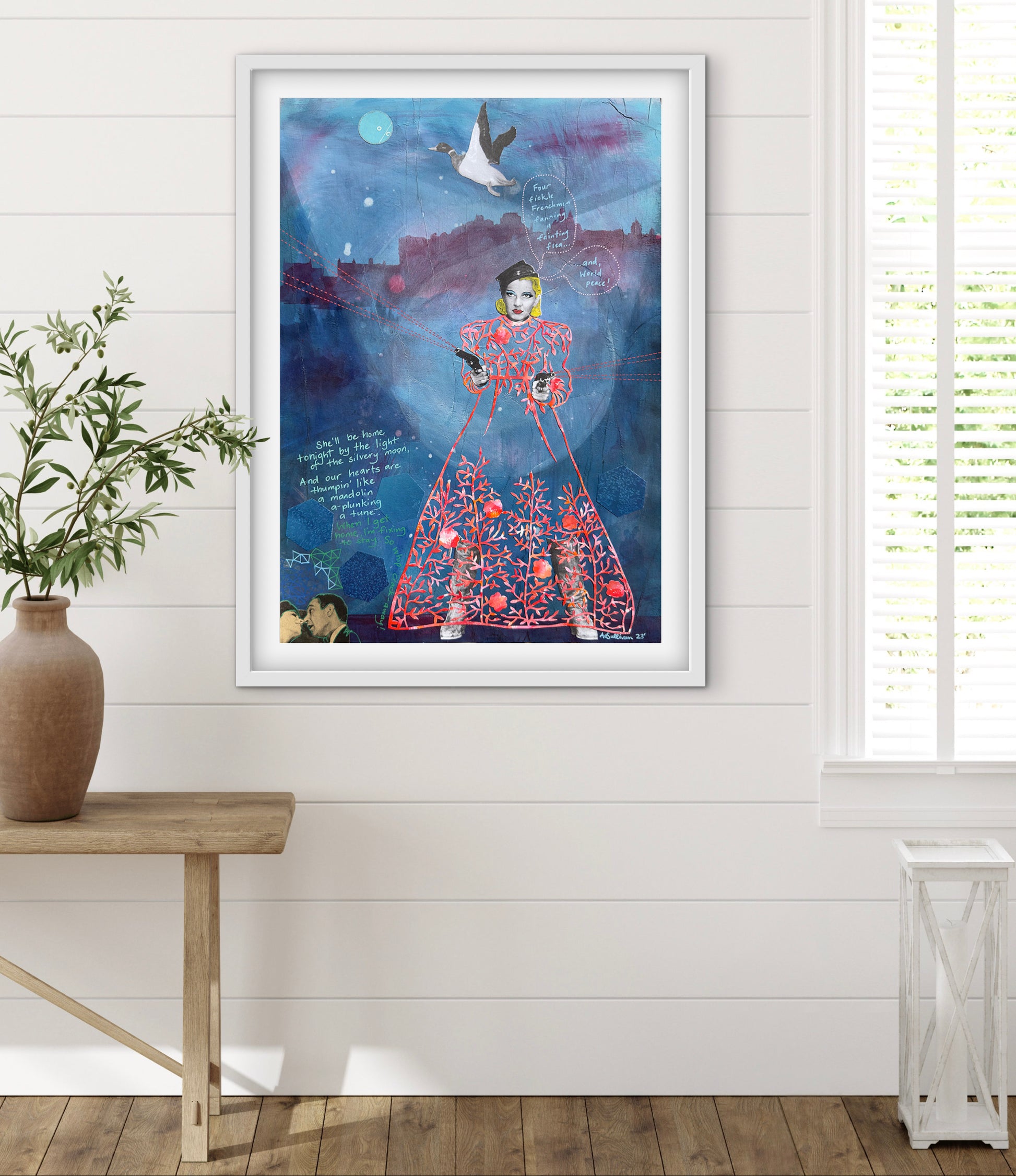 Framed artwork of a stylized figure against a blue background, displayed on a white wall with a wooden table and plant.